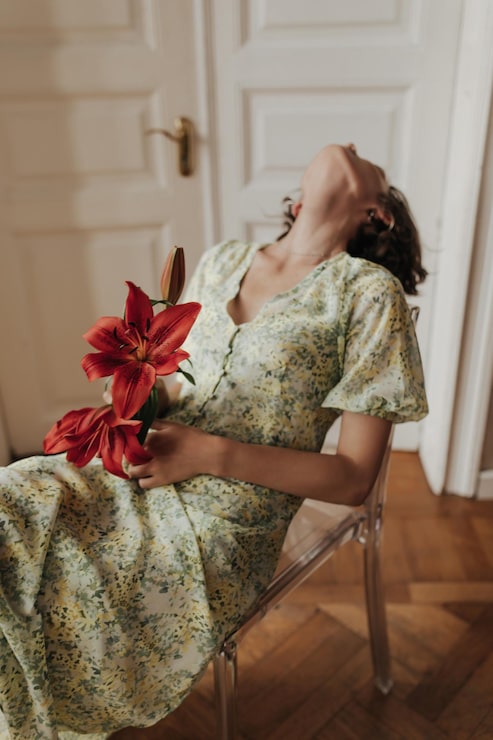 Foto gratuito sensible joven creativa emocional en midi floral vestido amarillo y verde se relaja en una silla transparente y sostiene flores rojas