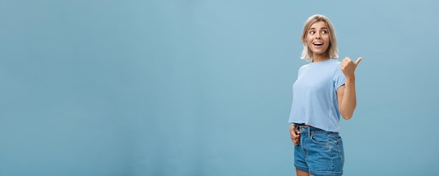 Free Photo charming sociable and friendly stylish female with blond hair turning right to show way or while discussing something talking with relaxed carefree look posing over blue background in trendy outfit