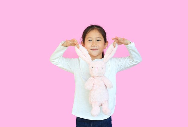 Photo เด็กหญิงชาวเอเชียตัวน้อยน่ารัก ถือหูกระต่ายสีชมพู แยกจากพื้นหลังสีชมพู เด็กมีความสุขในวันอีสเตอร์