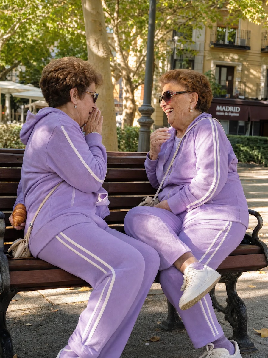 Dos abuelas con chándales lilas a juego riendo en un banco de un parque en Madrid, una de ellas escondiendo un cruasán a la espalda, luz moteada de tarde